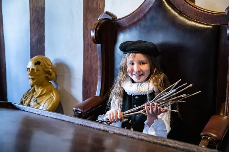 school pupil dressed in Tudor clothing stat on a desk chair smiling at Shakespeares schoolroom