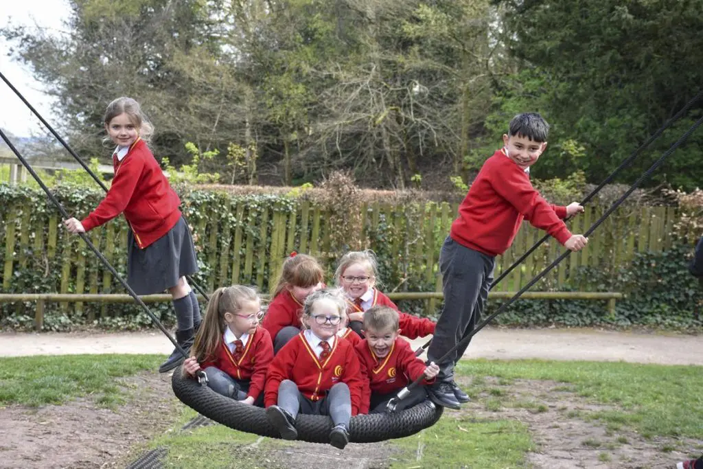 school children smiling and laughing on a large rope swing in a play area