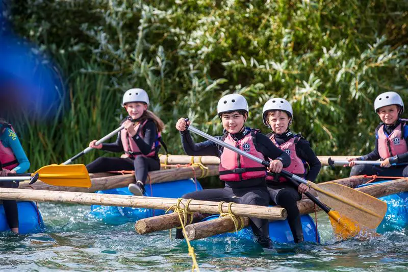 Willen Lake Day Trips Milton Keynes 6 school pupils on a raft paddling on a lake with wet suits, hard hats and life jackets on