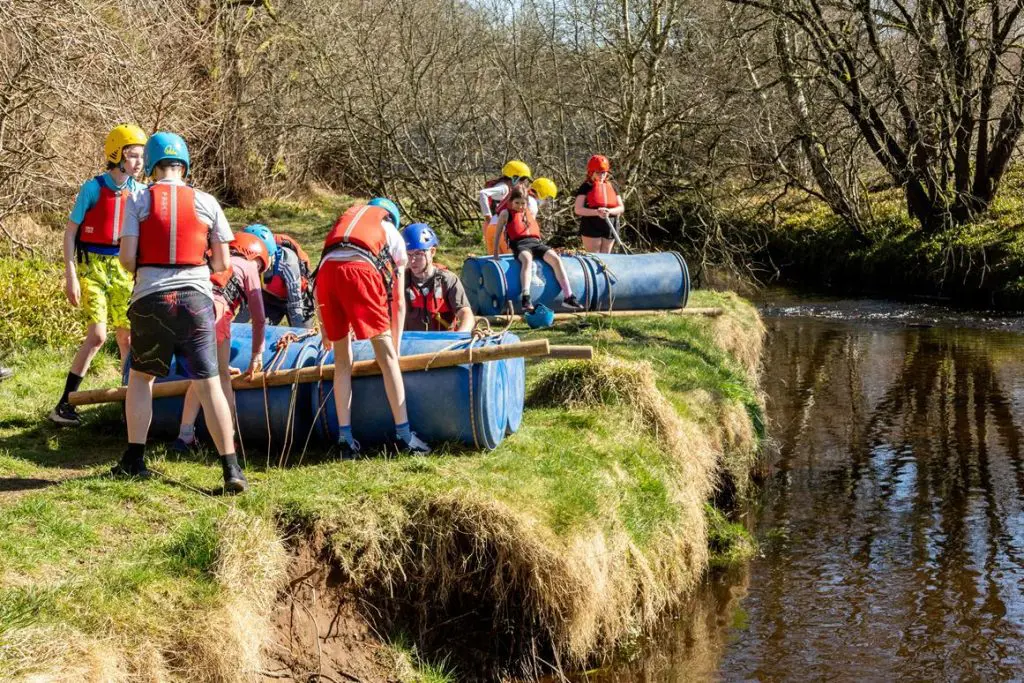 school trip group in harts had and life jackets by a river bank about to drop some man made rafts into the water