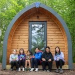 school group sitting by a wooden log cabin in a forest area