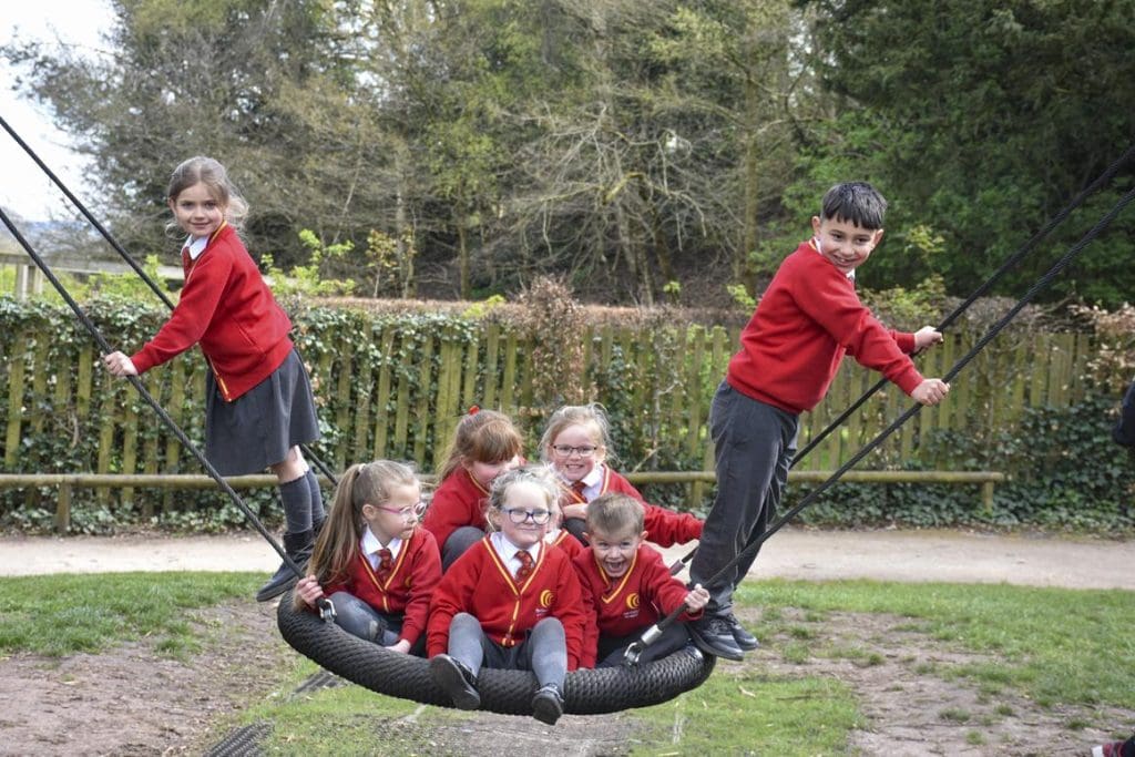 school children smiling and laughing on a large rope swing in a play area