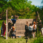 primary school students on a wooden climbing wall in a field by woods