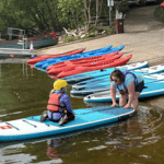 school pupil on paddle board being pushed out on lake by instructor