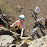 group of secondary school pupils with hard hats and safety gear climbing with saftey ropes up a rocky cliff edge
