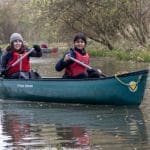 two secondary school students in a canoe with paddles on a river with safety gear smiling