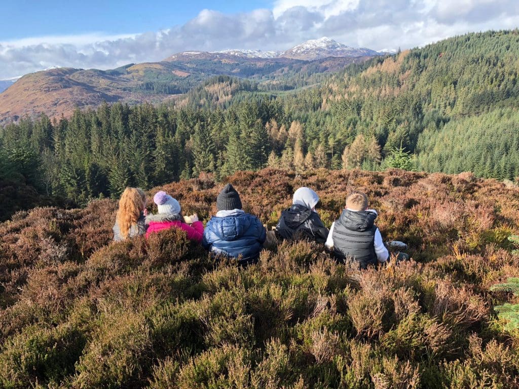 school trip group sitting on top of a hill outside surrounded with an amazing view of trees and distant slow lying mountain in Scotland