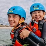 primary school pupils in the water with wet suits helmets and life jackets on smiling and having fun