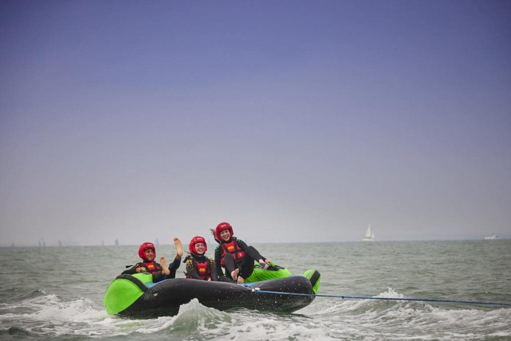 three school pupils in wet gear on a green inflatable on the sea being towed along by speed boat